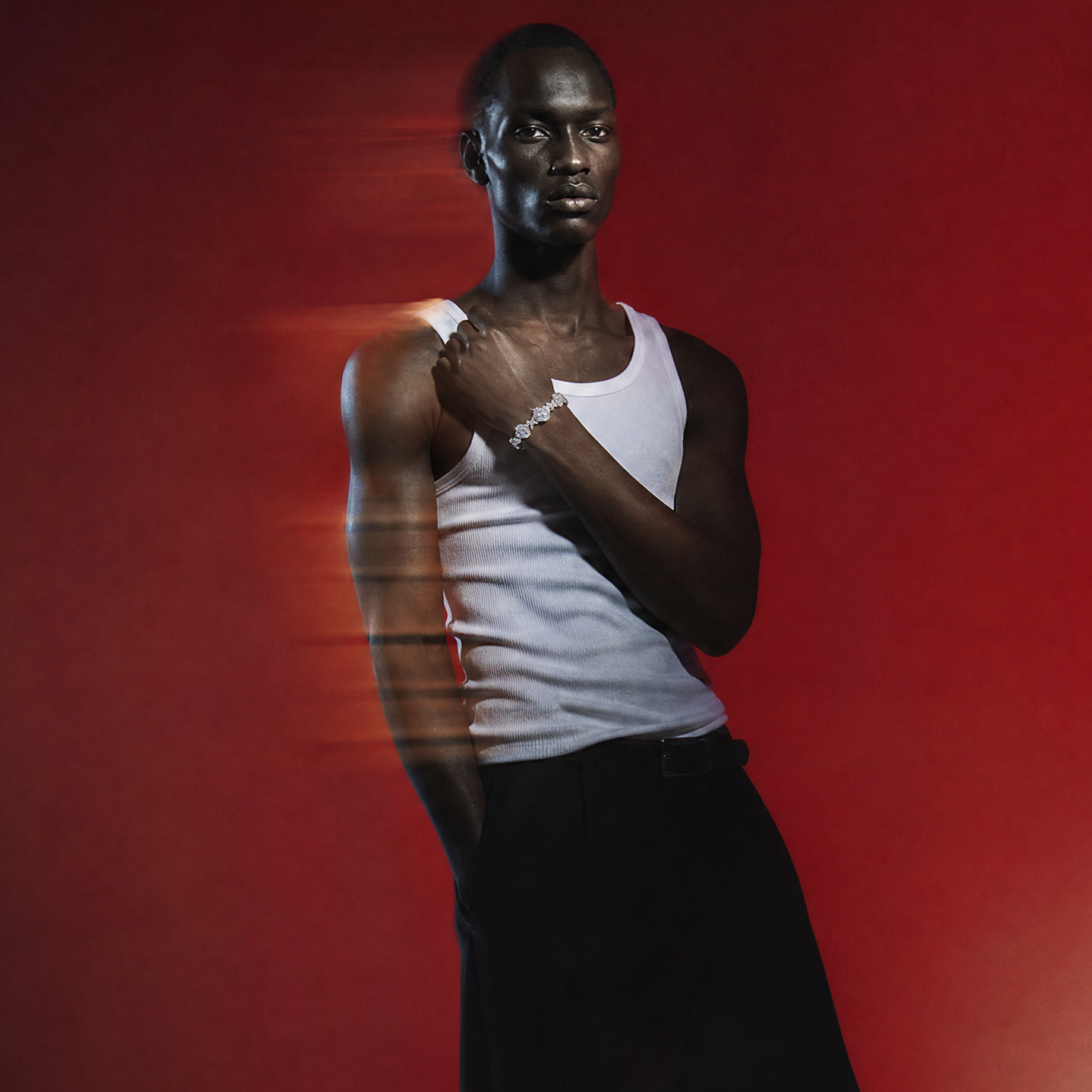 A man stands in a studio wearing a white tank top and loose black trousers against a red gradient background with motion blur streaks across his body.